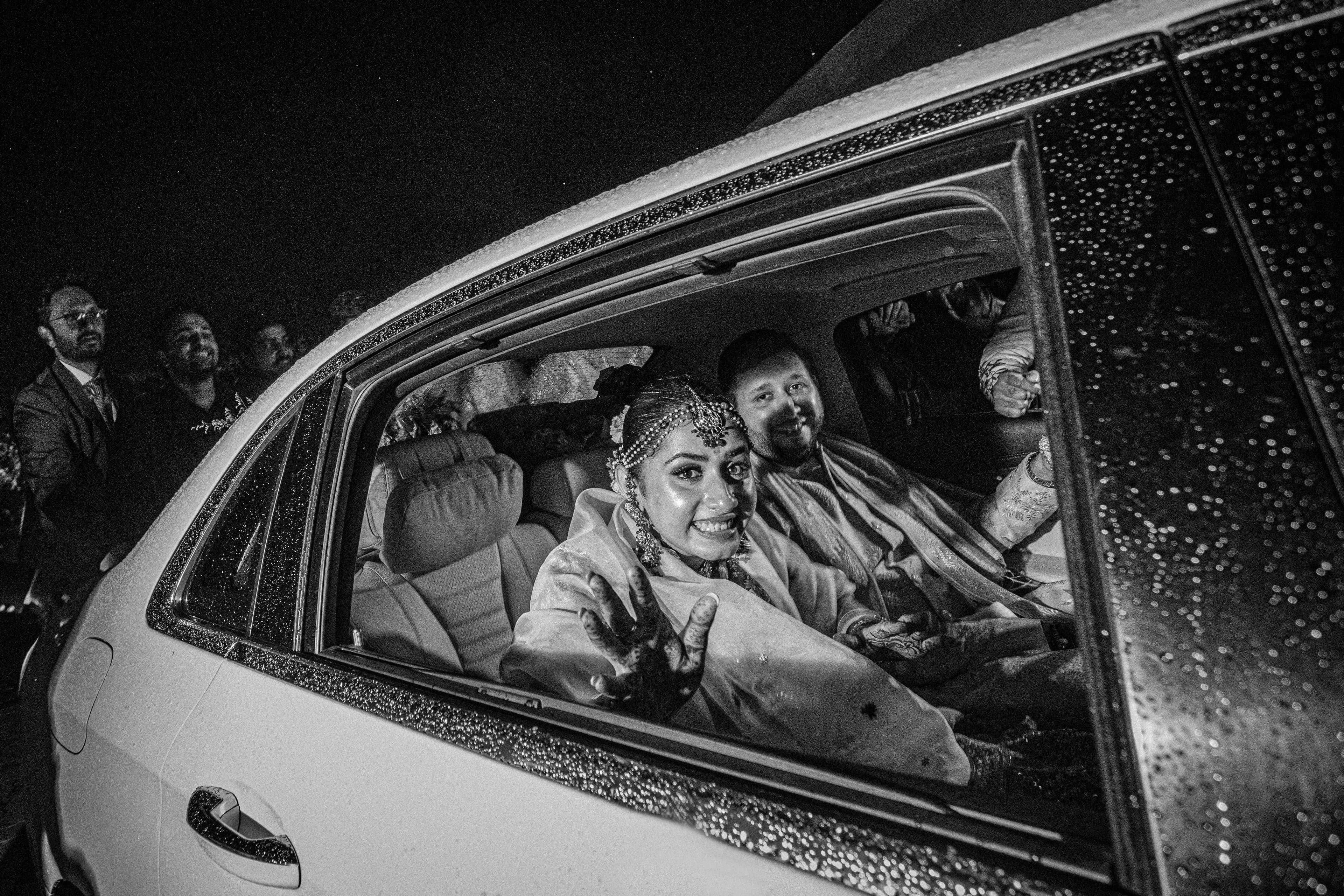 Bride and groom smiling from the back seat of a car at night, rain on the window
