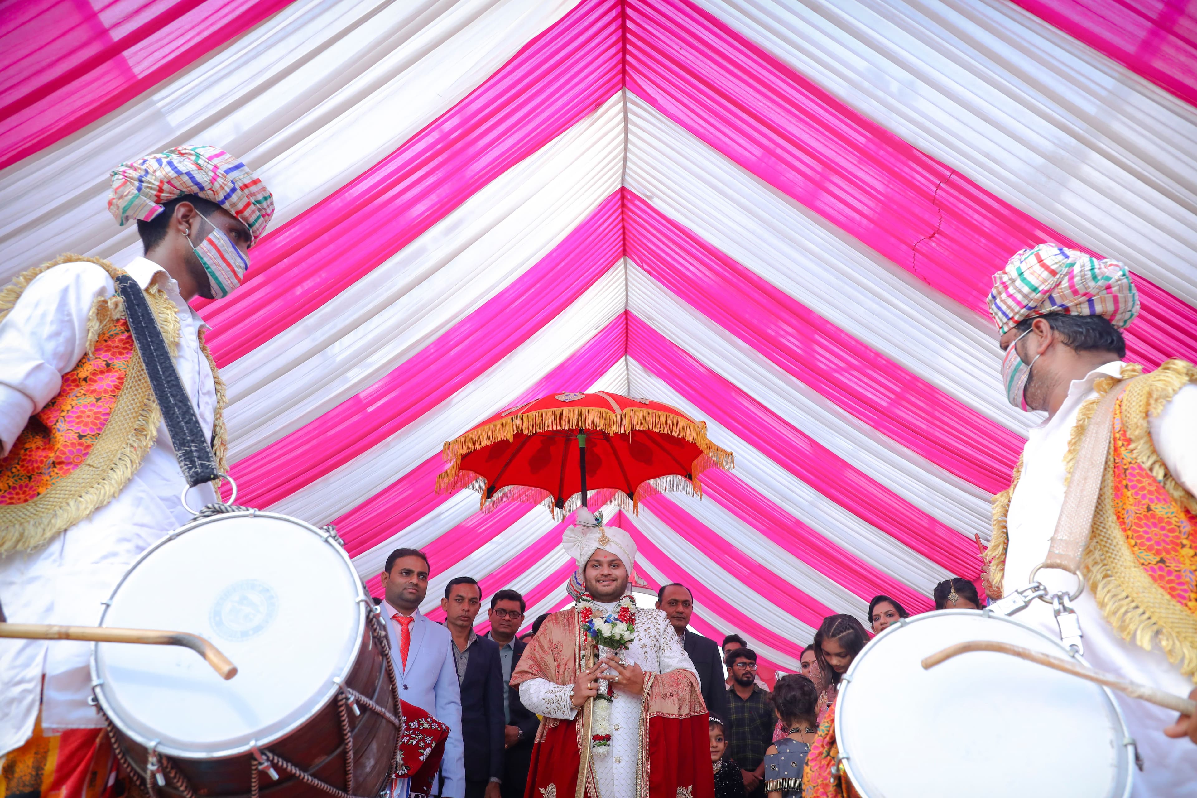Groom walking under a red umbrella flanked by two dhol players in colorful turbans